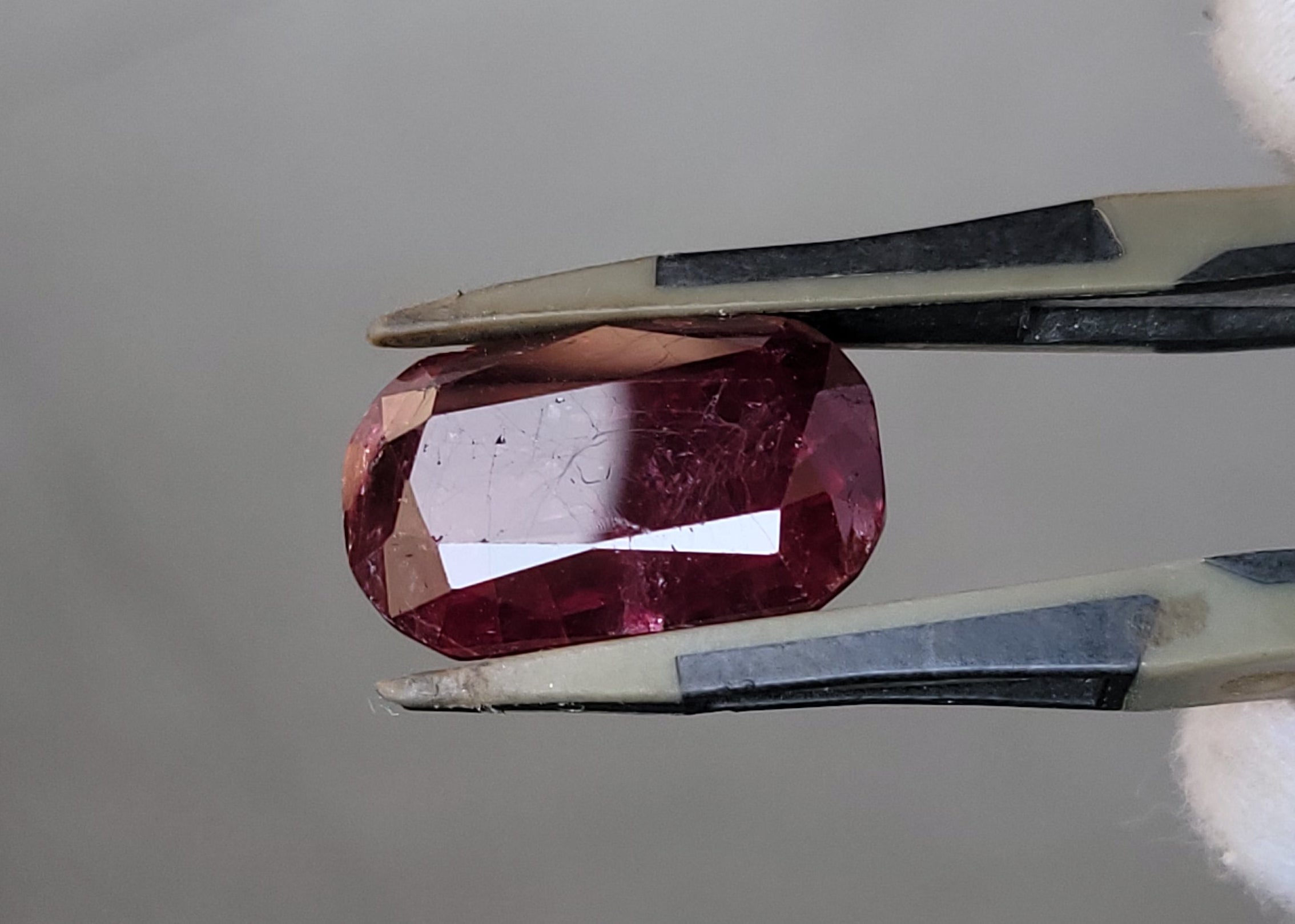 A faceted oval pink gemstone held in silver metal tweezers against a soft gray background.