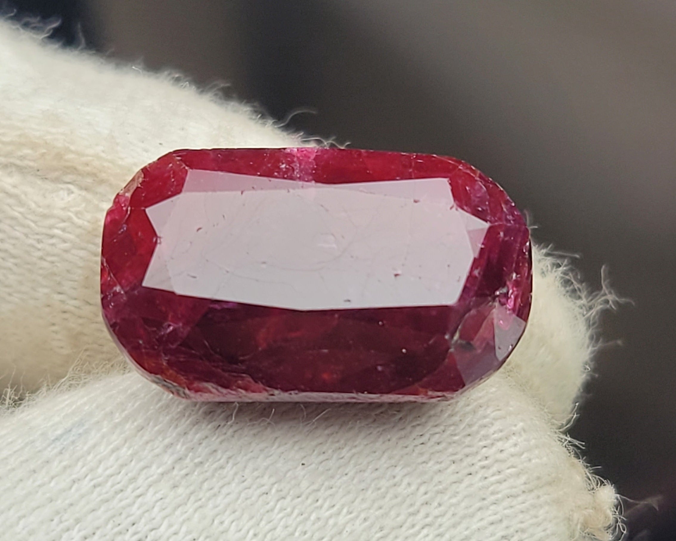 A faceted oval red gemstone held on a white knit gloved finger, photographed close-up against a blurred dark background.