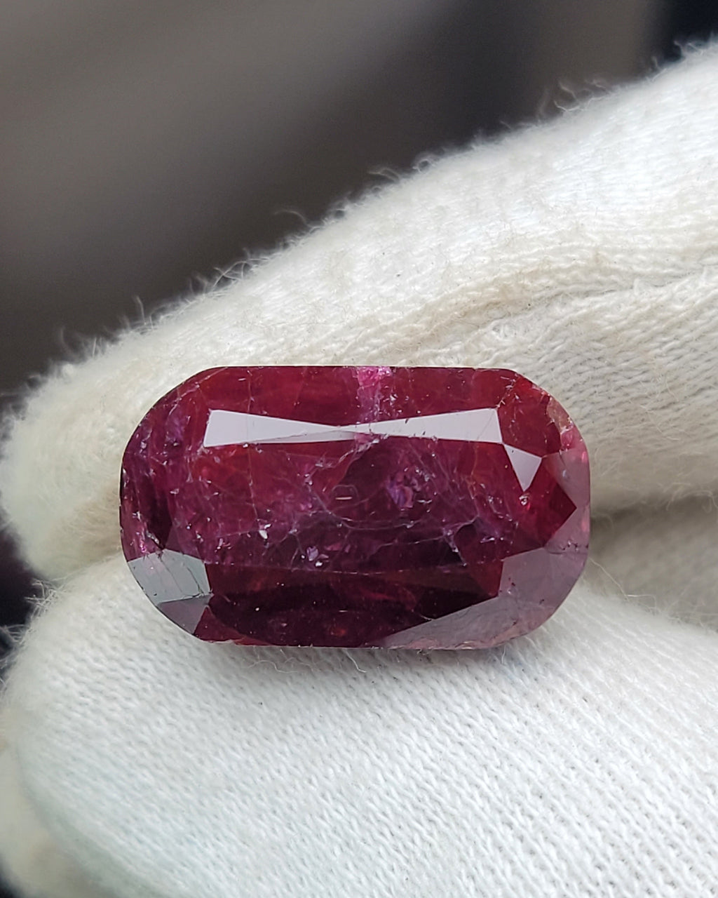 A cushion-cut deep pinkish-red gemstone held on a white textured glove.