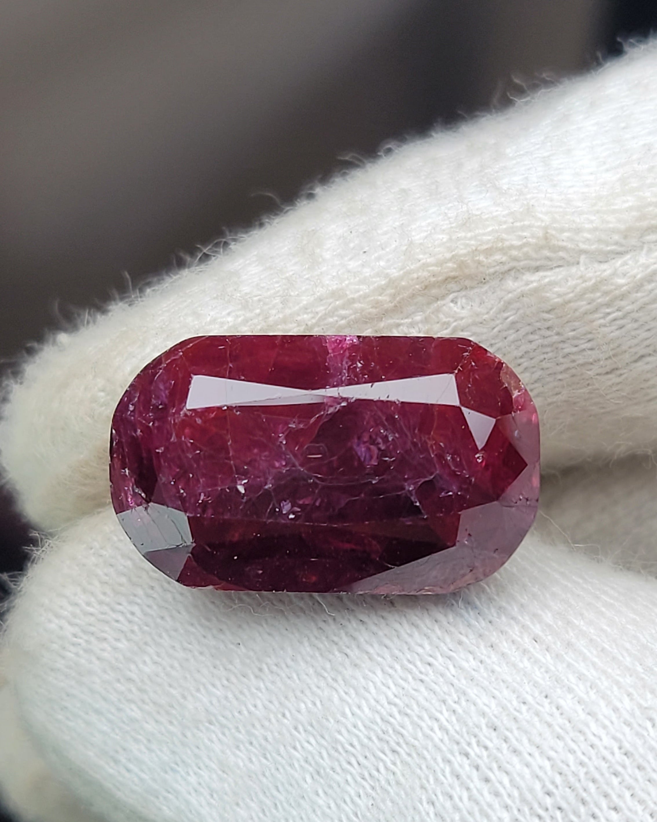 A cushion-cut deep pinkish-red gemstone held on a white textured glove.
