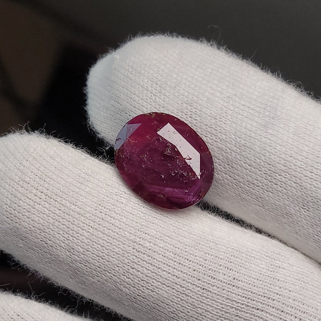 A round cabochon ruby gemstone with a deep magenta-pink hue, held on a white gloved finger against a dark gray background.
