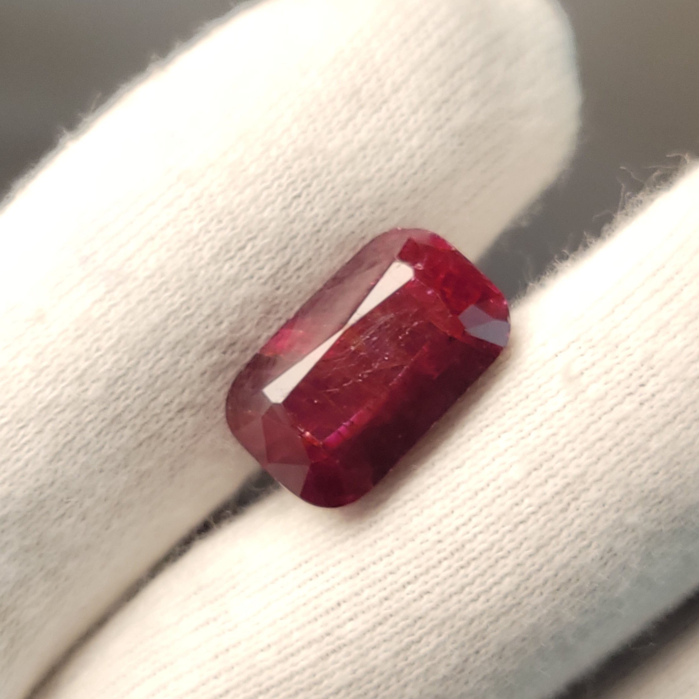 A rectangular emerald-cut deep red gemstone displayed on a white gloved finger.