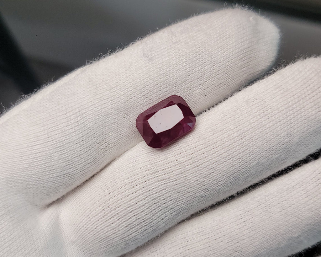 A small rectangular faceted ruby gemstone displayed on a white gloved finger, with a dark window background.