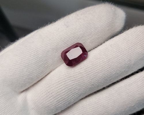 A small rectangular faceted ruby gemstone displayed on a white gloved finger, with a dark window background.