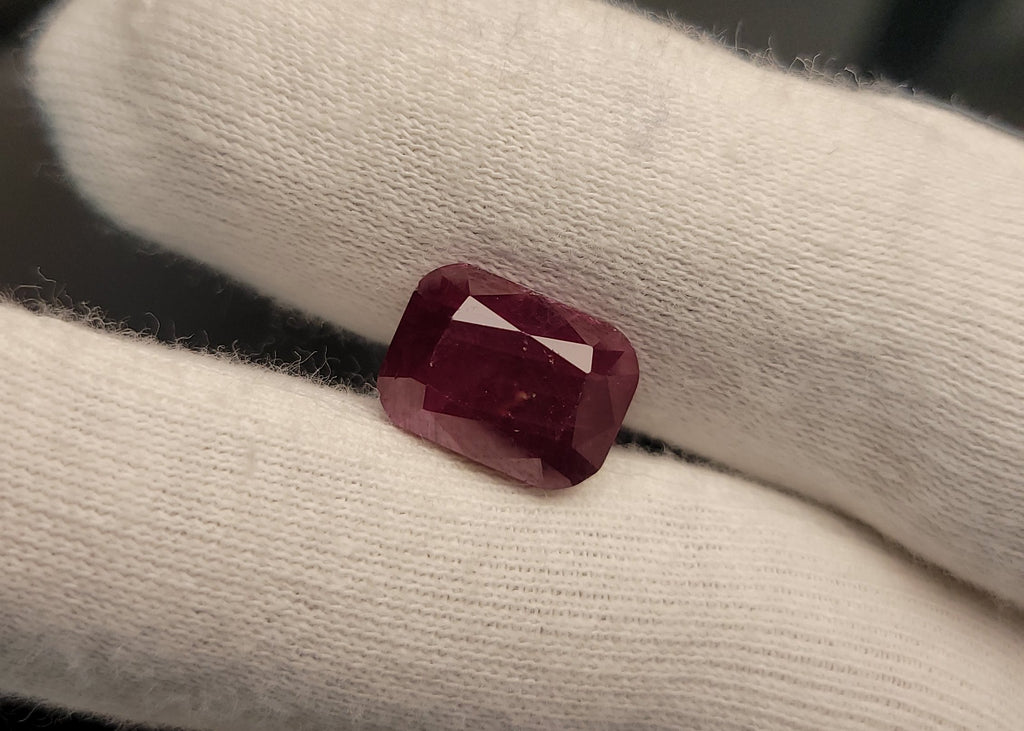 A rectangular cushion-cut deep red gemstone, likely a ruby, resting on a white gloved finger.