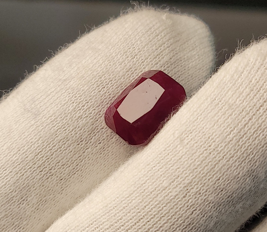 A small rectangular red gemstone with a white center, held between white gloved fingers.