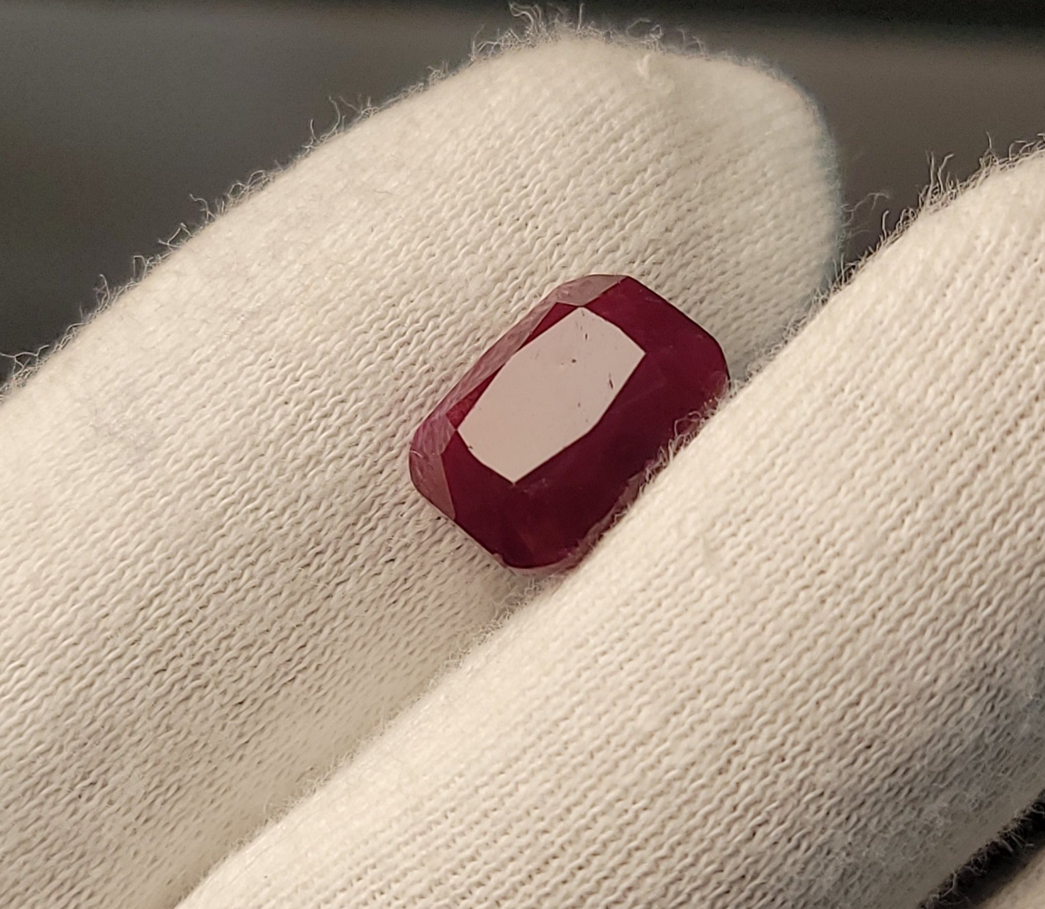A small rectangular red gemstone with a white center, held between white gloved fingers.