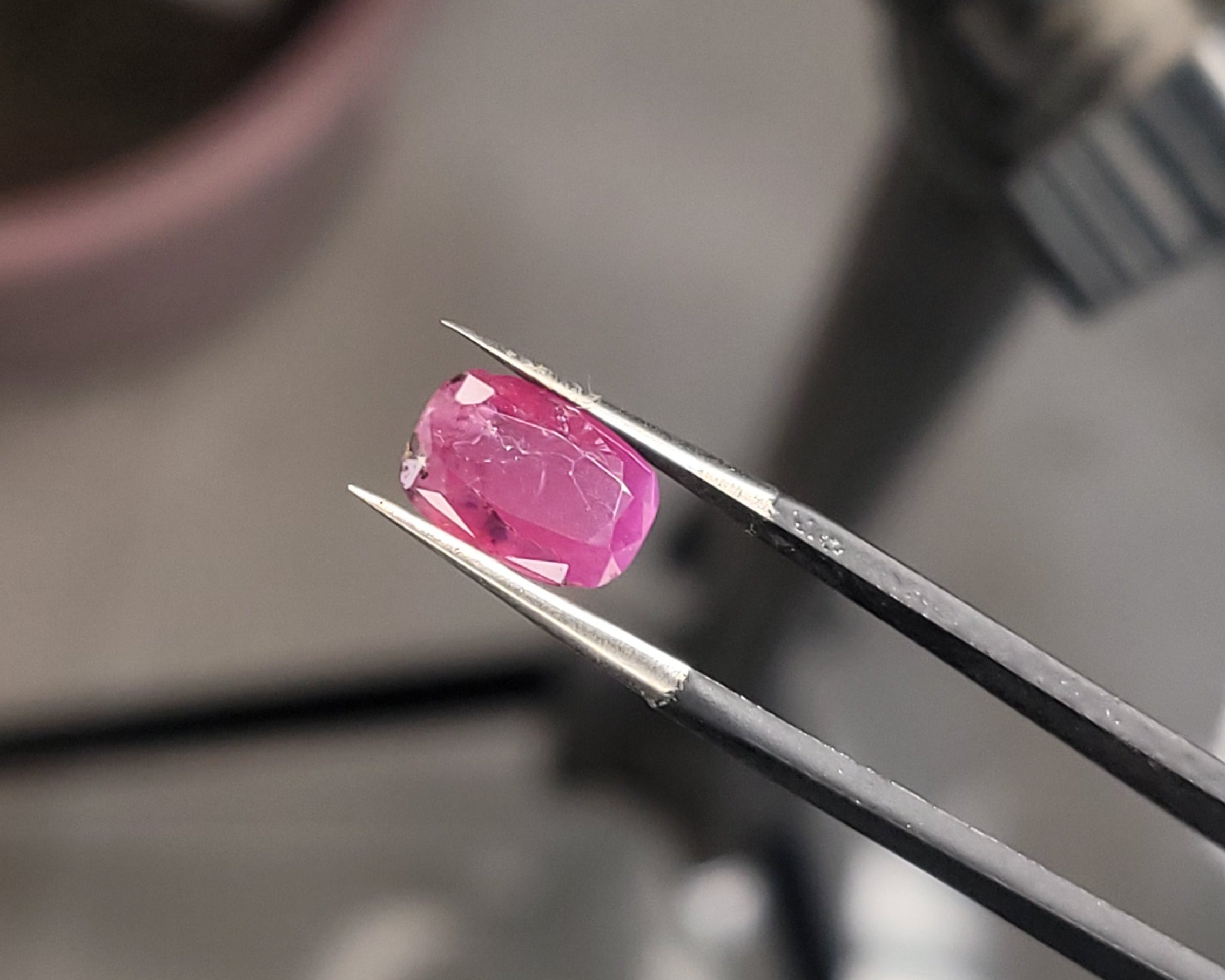 A small pink oval gemstone held between metal tweezers, photographed close up against a blurred workshop background.