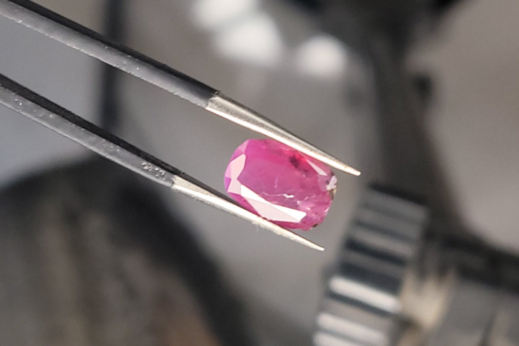 A small bright pink gemstone held in metal tweezers, photographed close up with a blurred workshop or studio background.