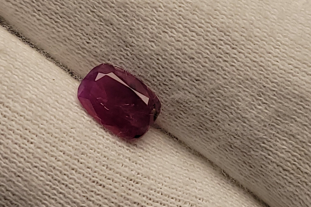 A small rectangular faceted ruby gemstone in a deep magenta-pink hue, resting on a textured off-white knit fabric.