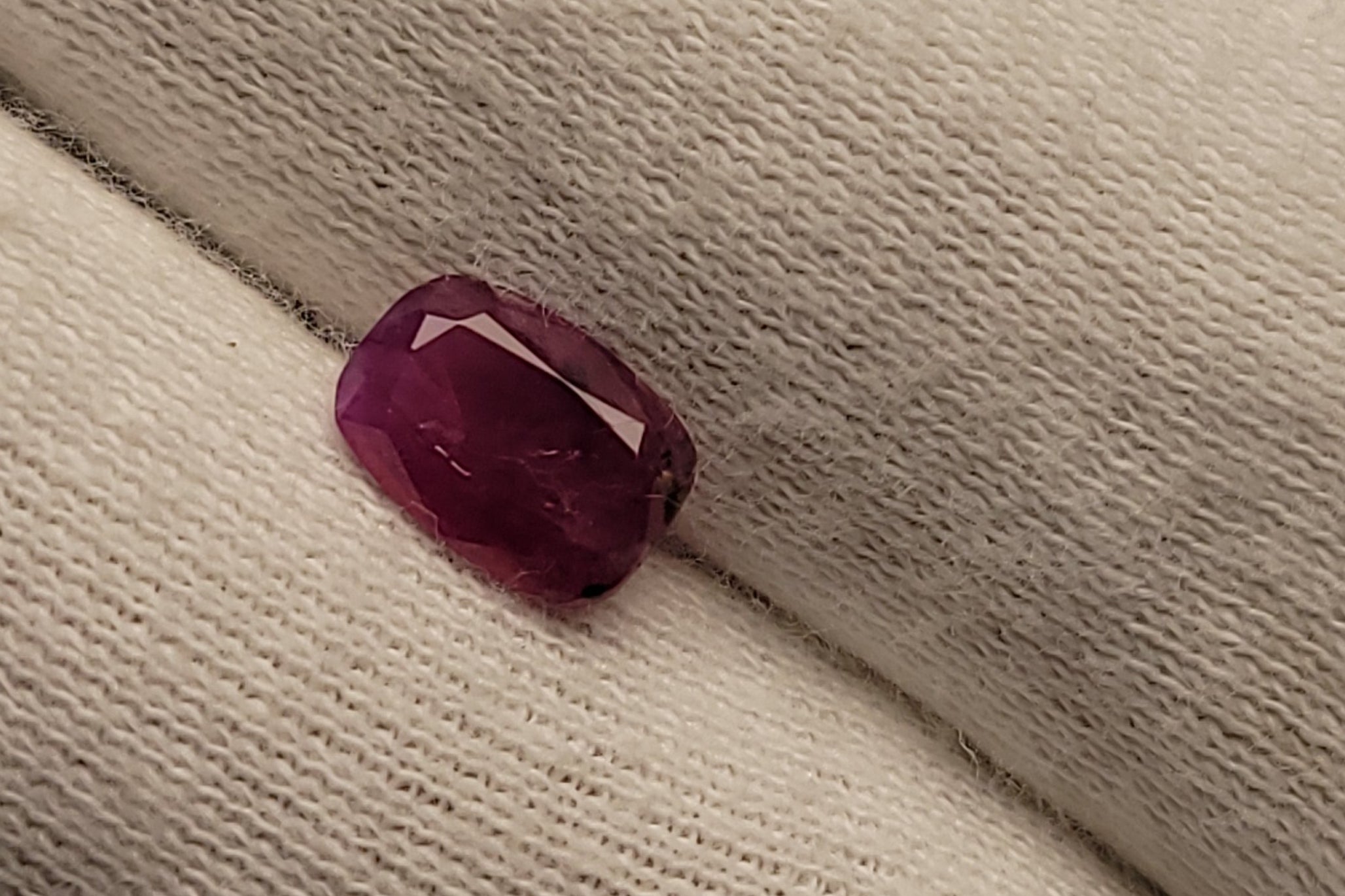 A small rectangular faceted ruby gemstone in a deep magenta-pink hue, resting on a textured off-white knit fabric.