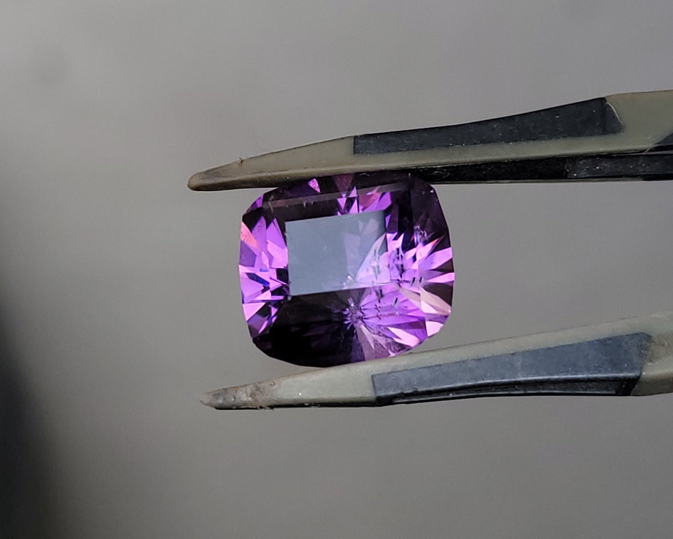 A cushion-cut purple gemstone held in silver metal tweezers against a plain light gray background.