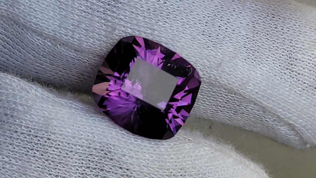A cushion-cut purple gemstone with a faceted, starburst-like internal pattern, resting on a white textured knit glove.