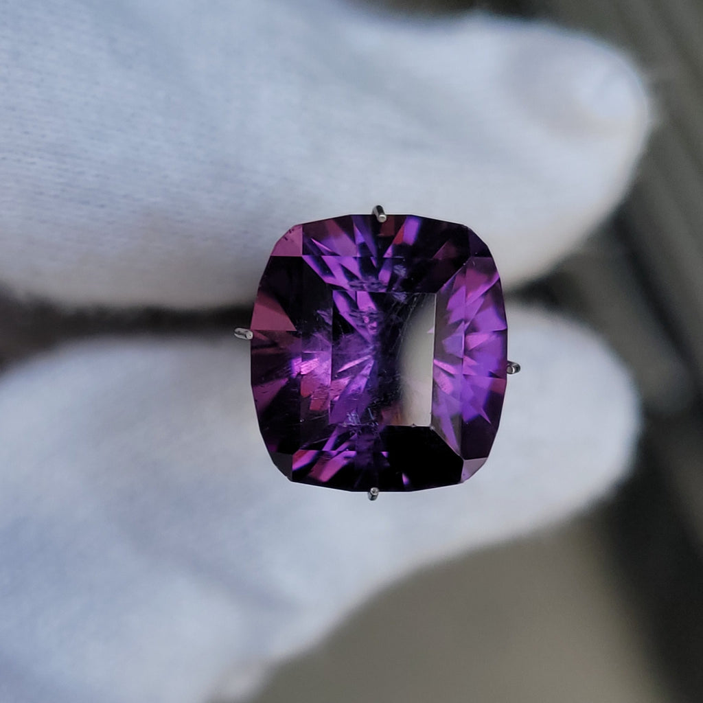 A cushion-cut purple gemstone with deep violet and magenta facets, held by a white gloved hand.
