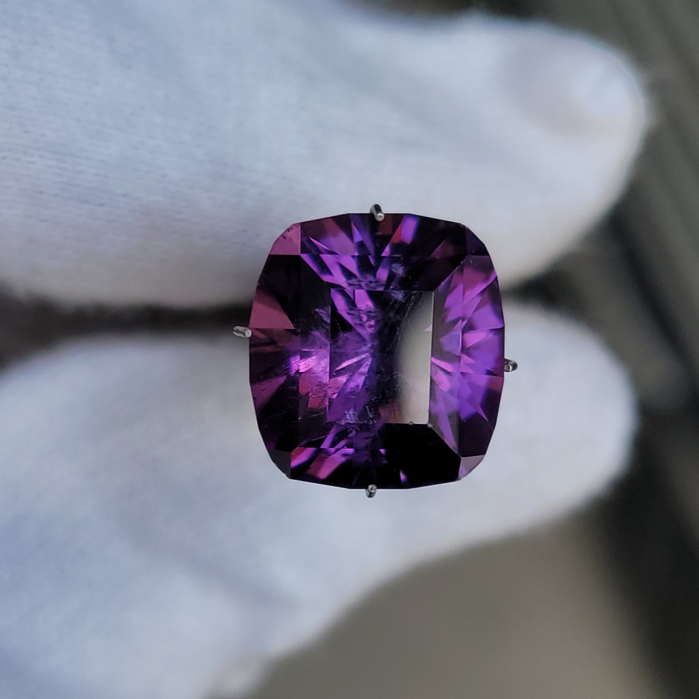 A cushion-cut purple gemstone with deep violet and magenta facets, held by a white gloved hand.