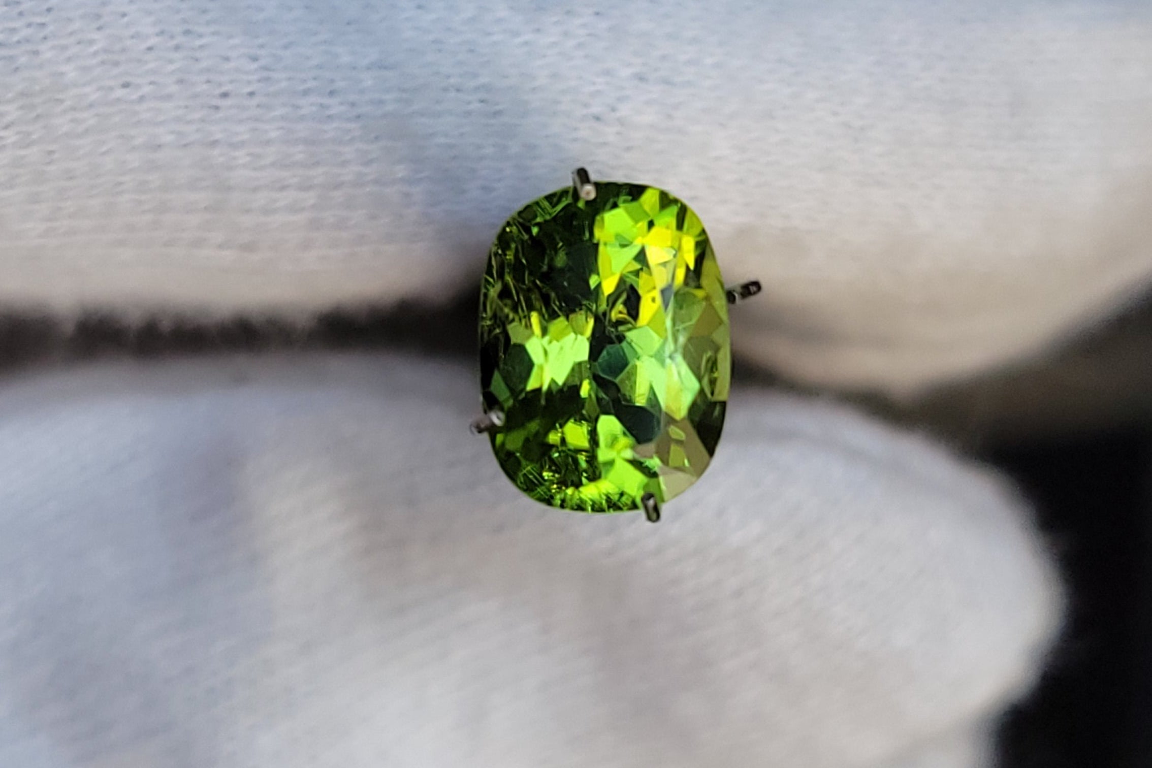 A vibrant green oval gemstone held between white gloved fingers against a dark, blurred background.