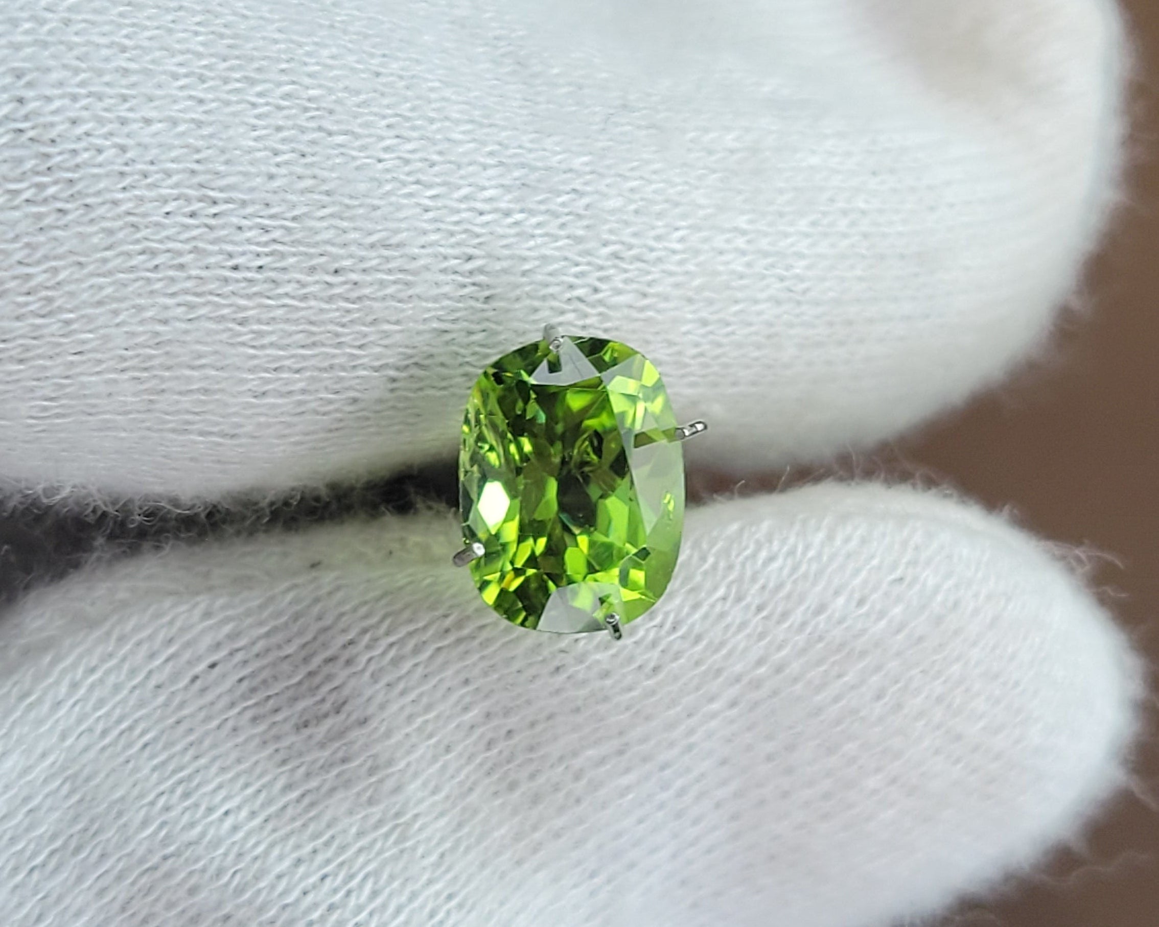 A small oval green gemstone held between white gloved fingers against a blurred brown background.