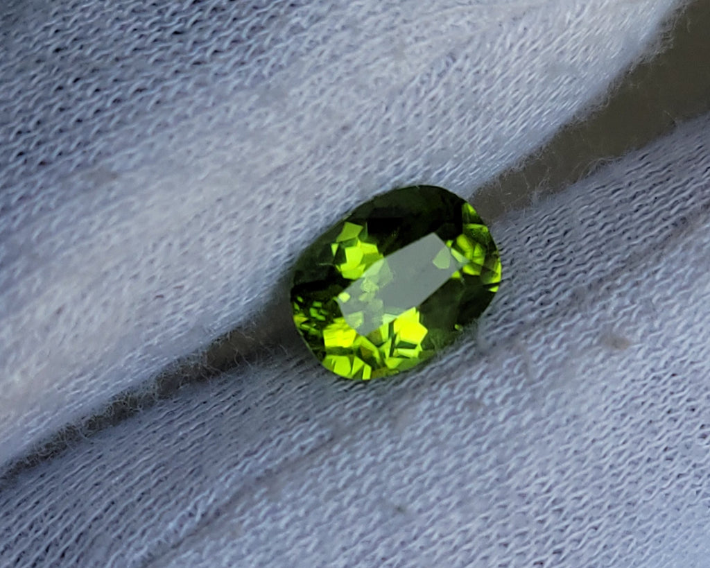 A small oval-cut green gemstone with bright yellow-green highlights, resting on a white textured fabric surface.
