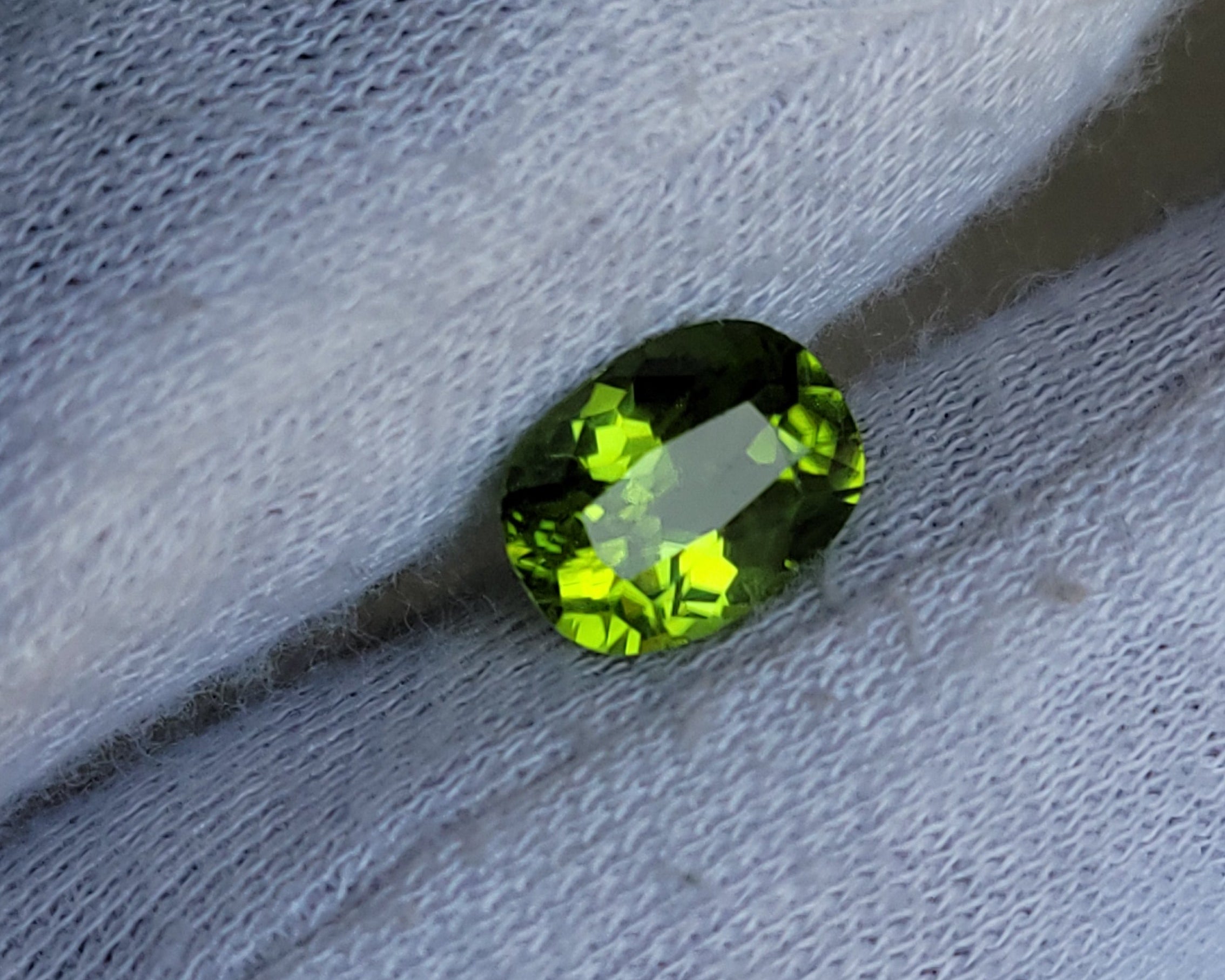 A small oval-cut green gemstone with bright yellow-green highlights, resting on a white textured fabric surface.