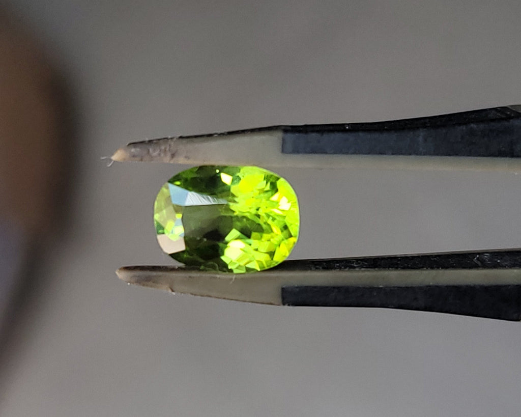 A bright yellow-green oval gemstone held between metal tweezers against a soft gray background.