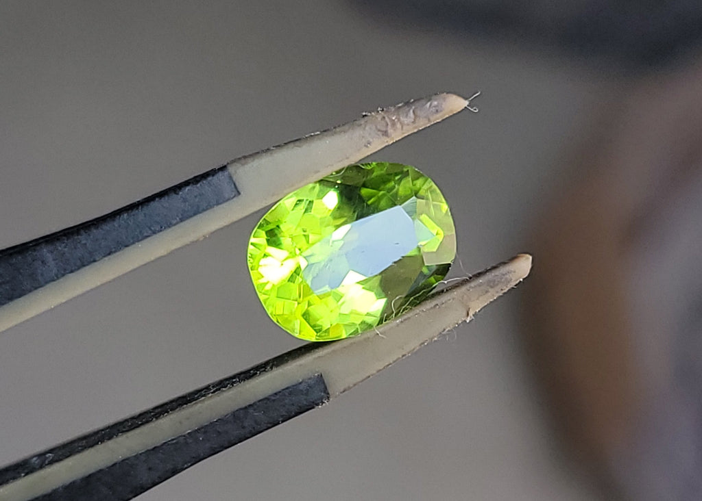 A bright green oval-cut gemstone held between metal tweezers against a softly blurred neutral background.