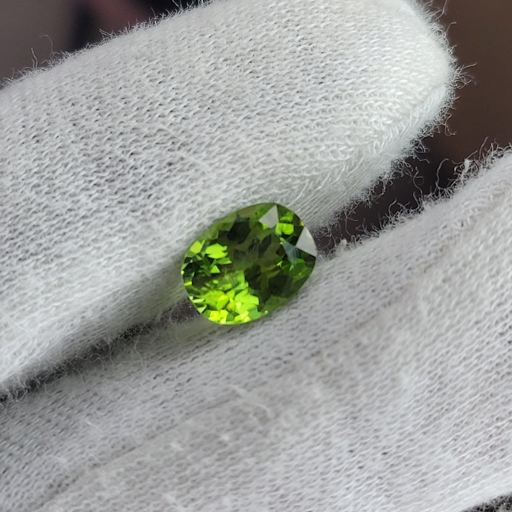 A small oval-cut bright green gemstone held between white textured gloves.