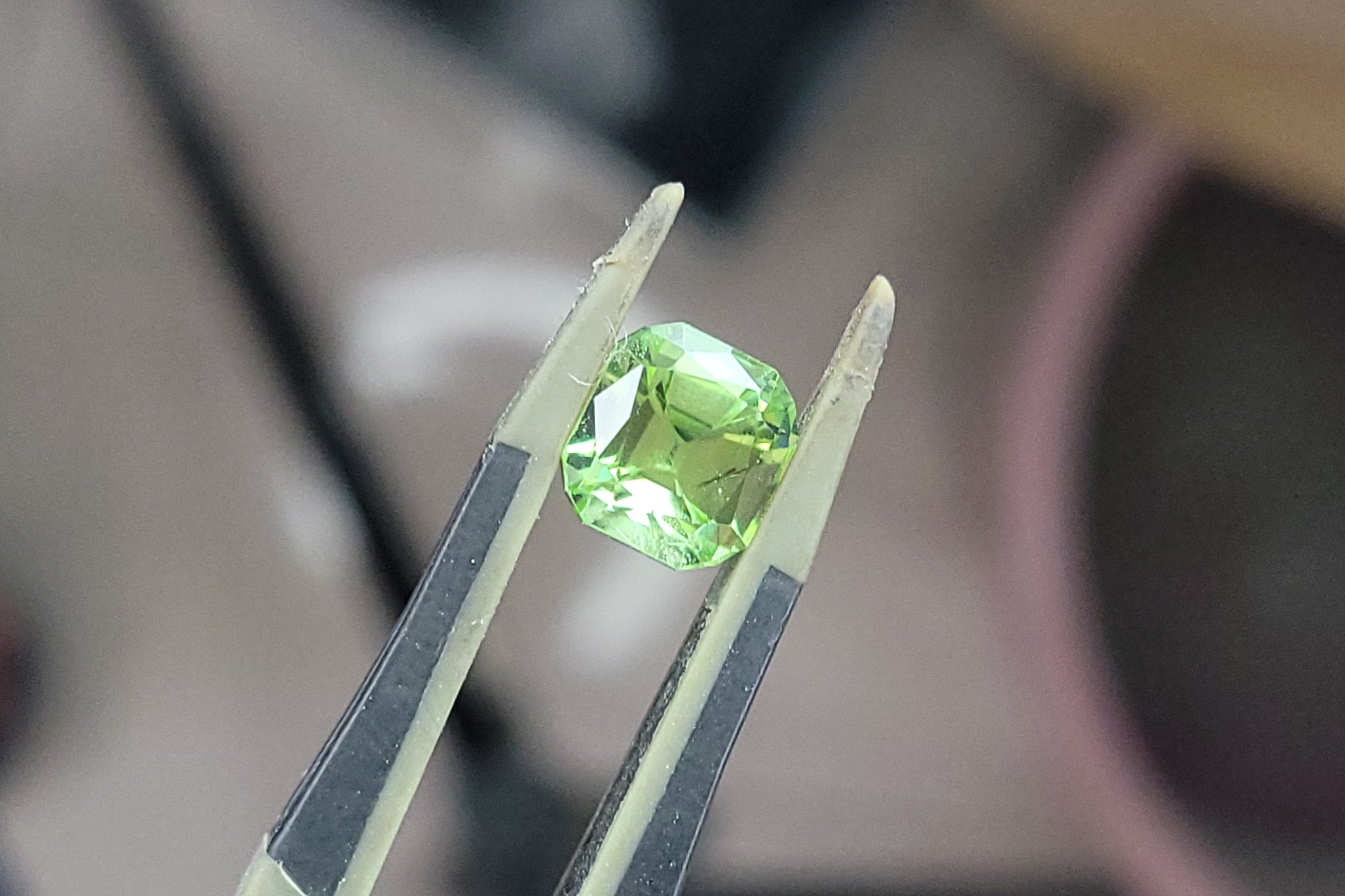 A small green faceted gemstone held between metal tweezers, photographed close up against a blurred workshop background.