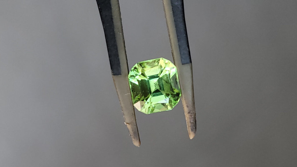 A faceted green gemstone held in a pair of metal tweezers against a plain gray background.