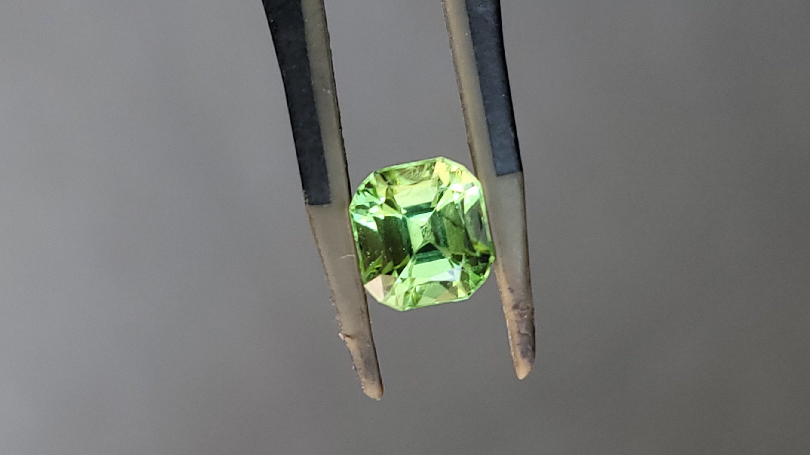 A faceted green gemstone held in a pair of metal tweezers against a plain gray background.