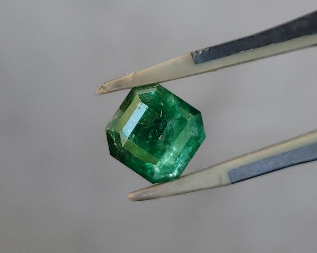 A faceted green gemstone held between metal tweezers against a soft, out-of-focus light gray background.