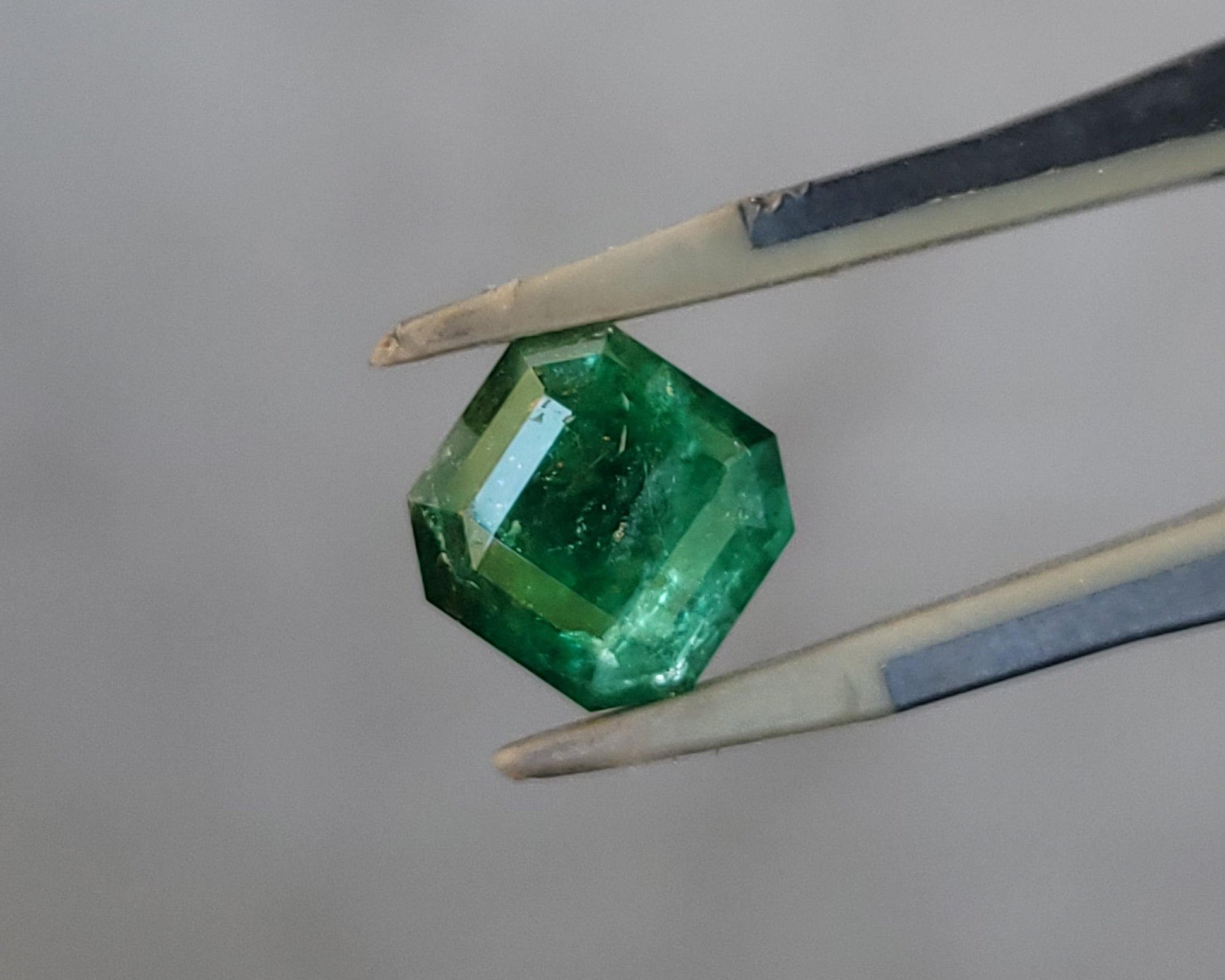 A faceted green gemstone held between metal tweezers against a soft, out-of-focus light gray background.