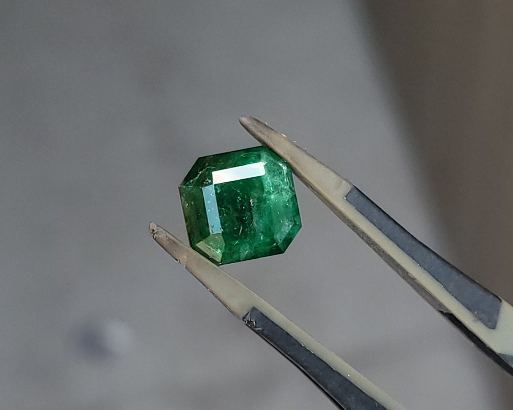 A small green faceted gemstone held between metal tweezers against a soft gray background.