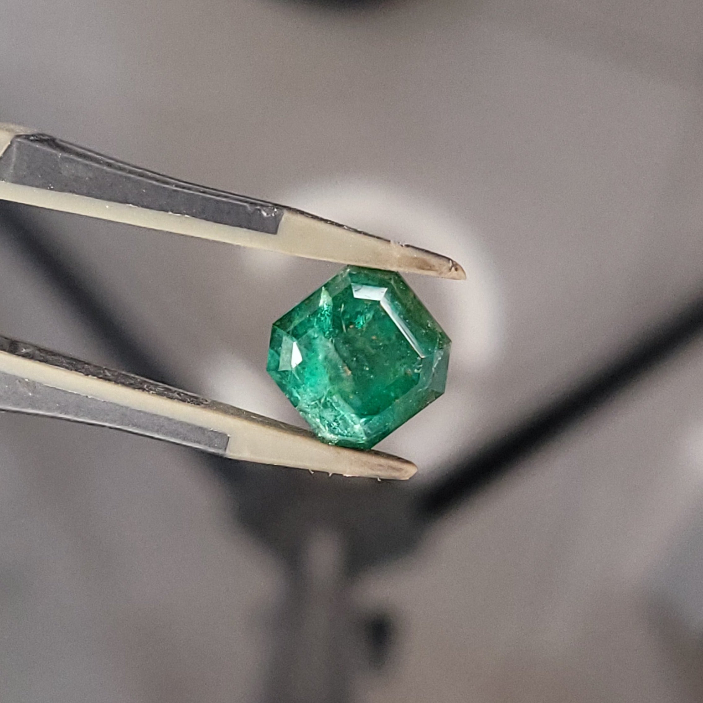 A green faceted gemstone held by metal tweezers in sharp focus, with a blurred workshop or studio background and a metallic stand visible.