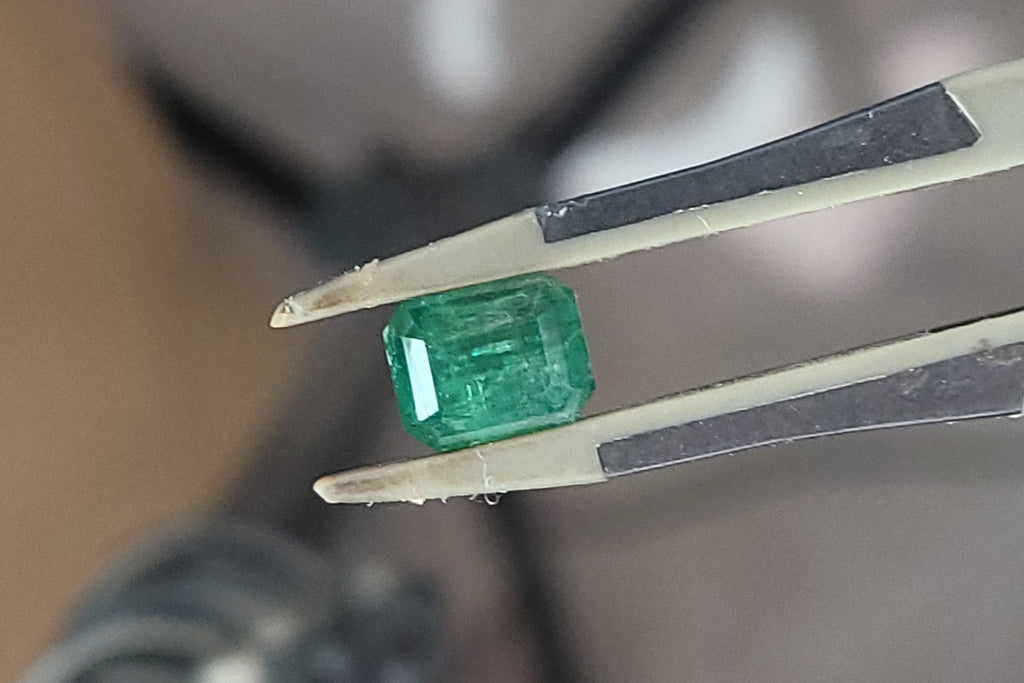 A small rectangular green gemstone held between metal tweezers, photographed close up with a blurred workshop or studio background.