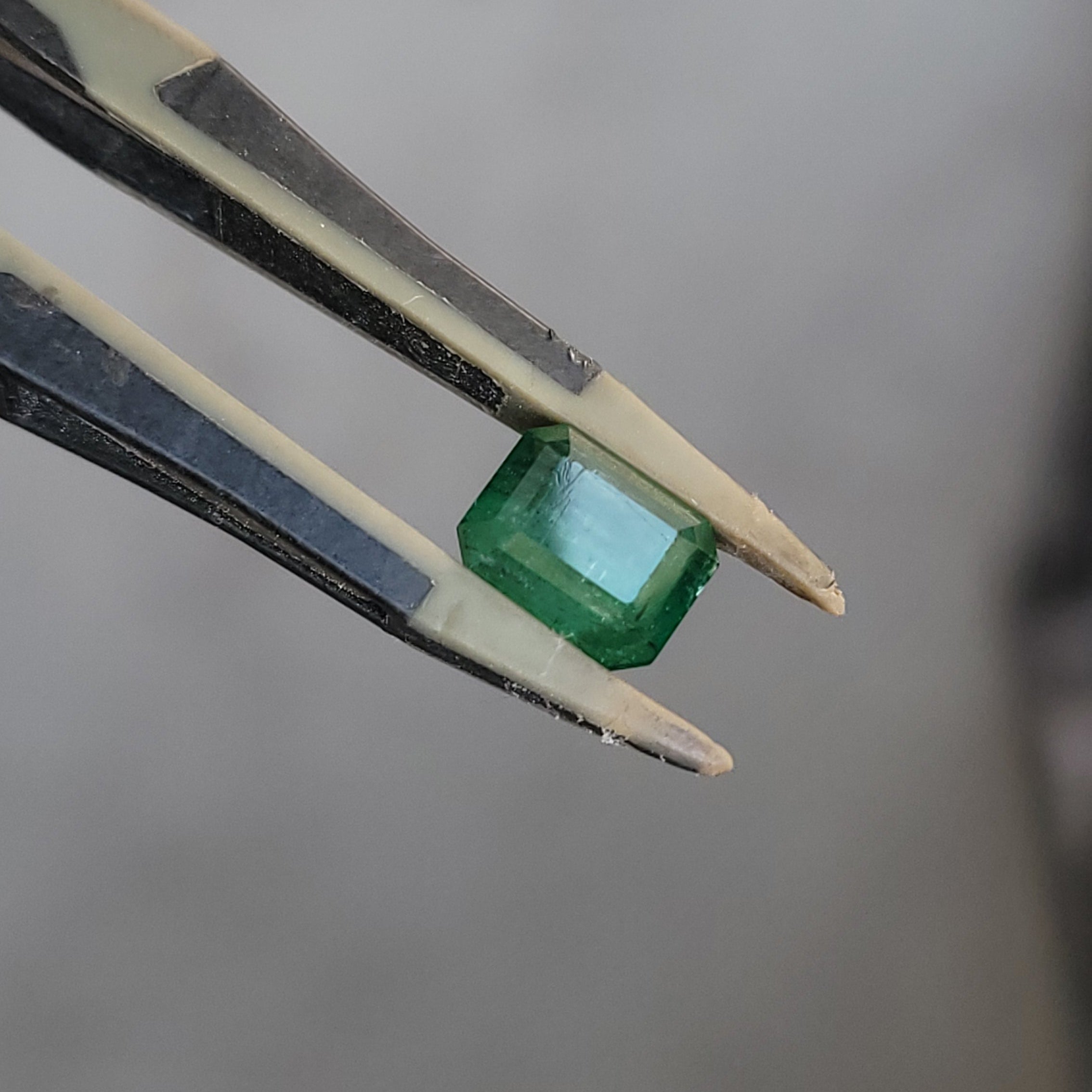 A green rectangular-cut gemstone held between metal tweezers against a blurred gray background.
