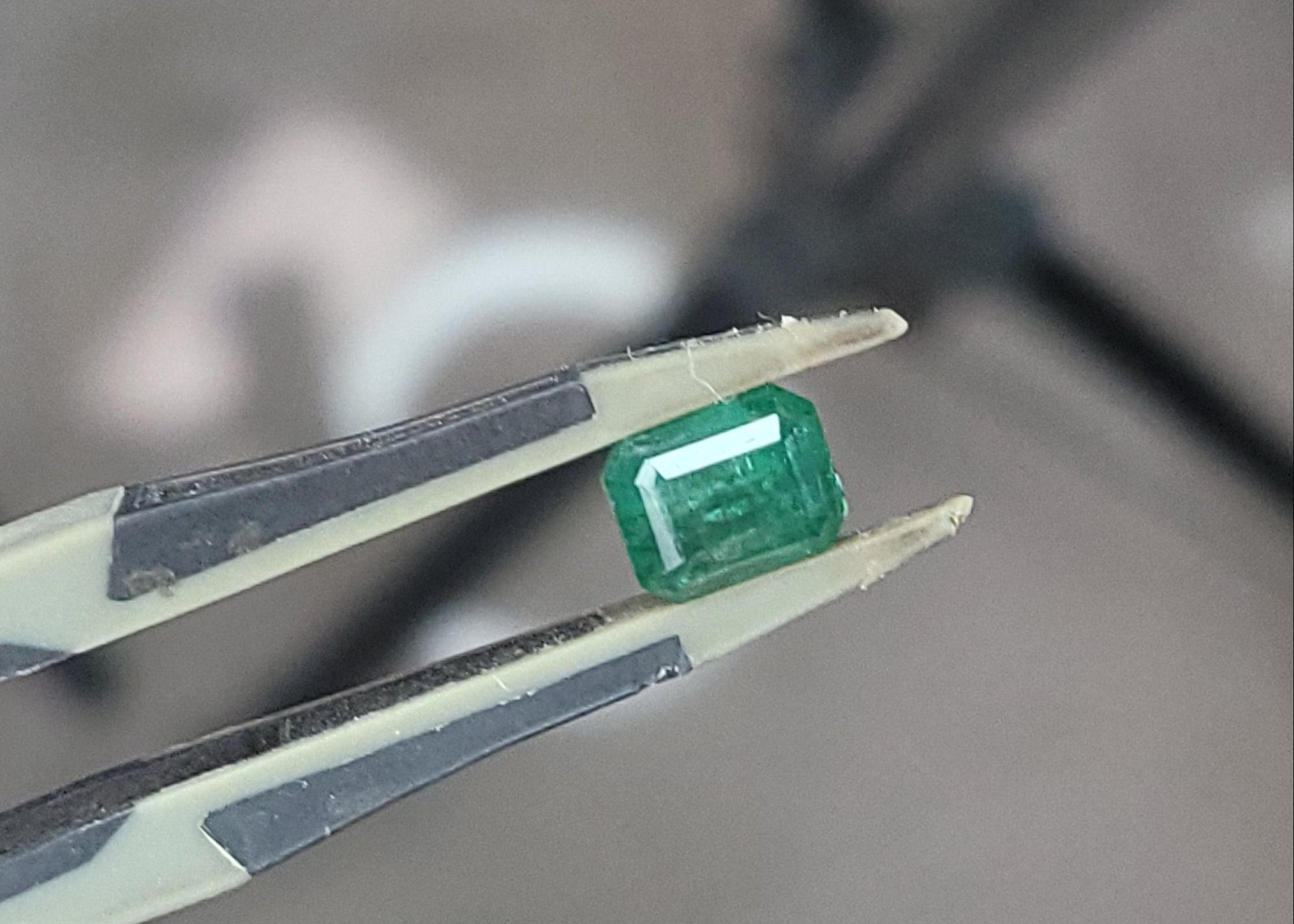 A small rectangular green gemstone held between metal tweezers, photographed in a real workshop or lab setting with blurred equipment and a foot visible in the background.