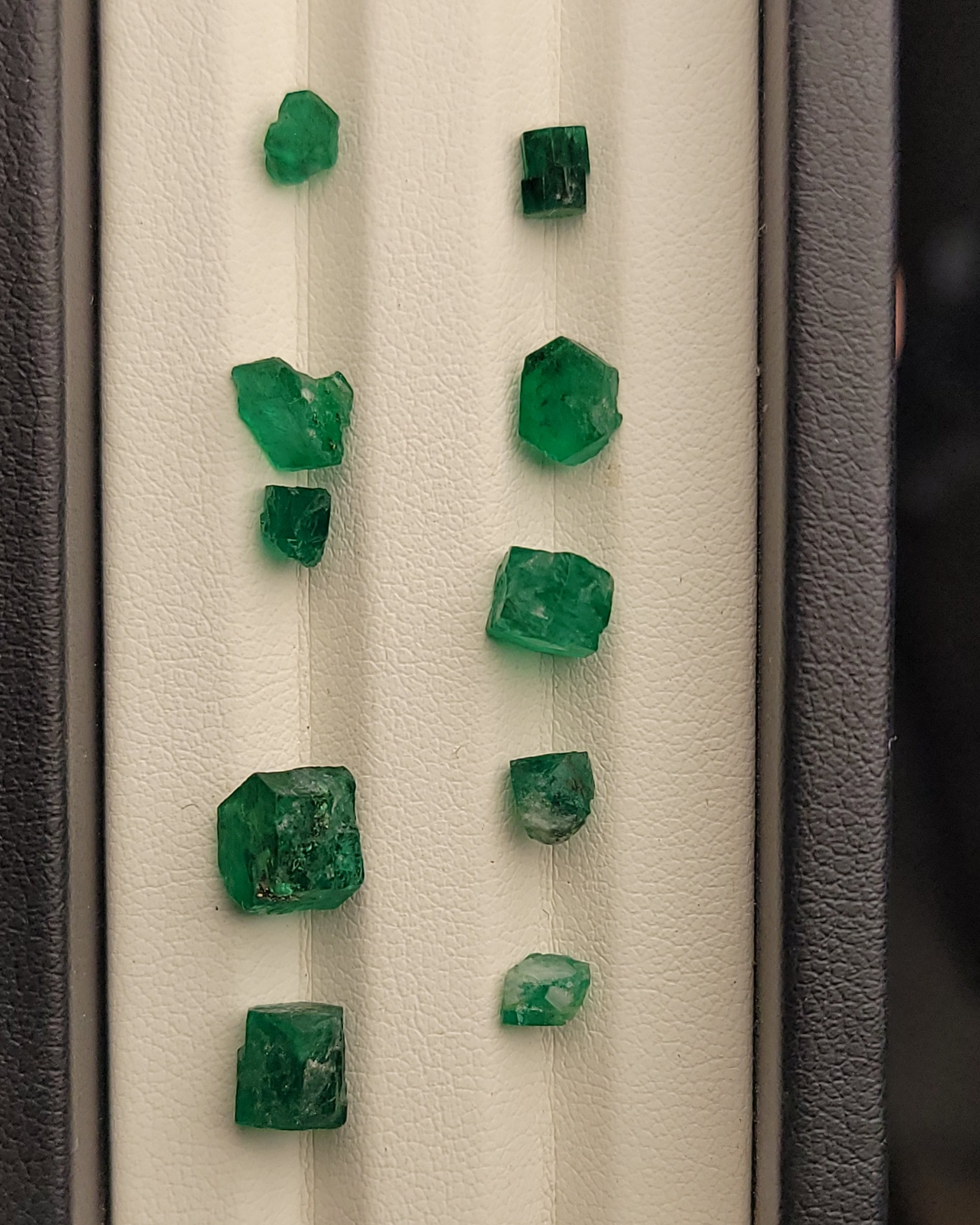 Several raw green emerald-cut gemstones arranged in two columns on a white textured display card inside a dark gray holder.