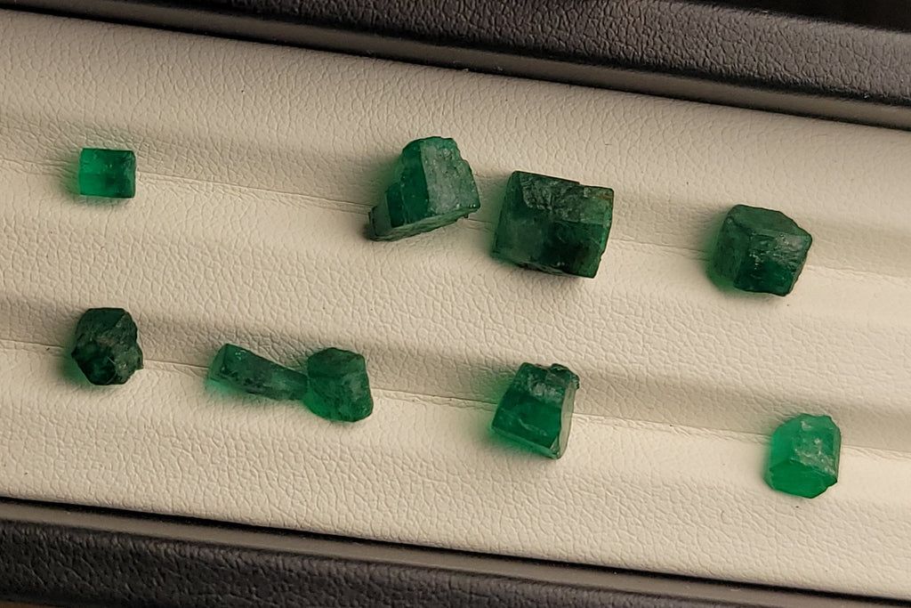 Several small green gemstone cubes displayed on a white cushion inside a black jewelry box, with a finger visible at the bottom edge.