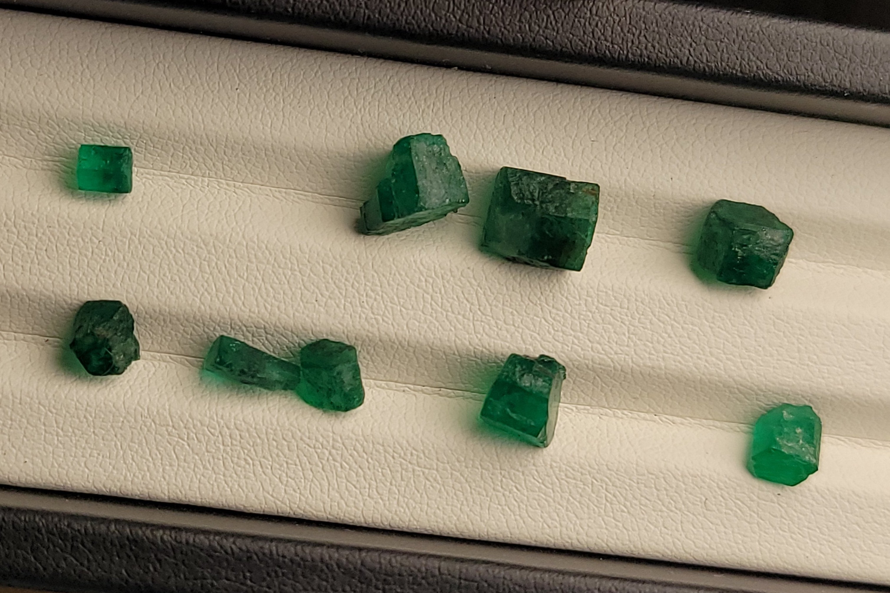 Several small green gemstone cubes displayed on a white cushion inside a black jewelry box, with a finger visible at the bottom edge.
