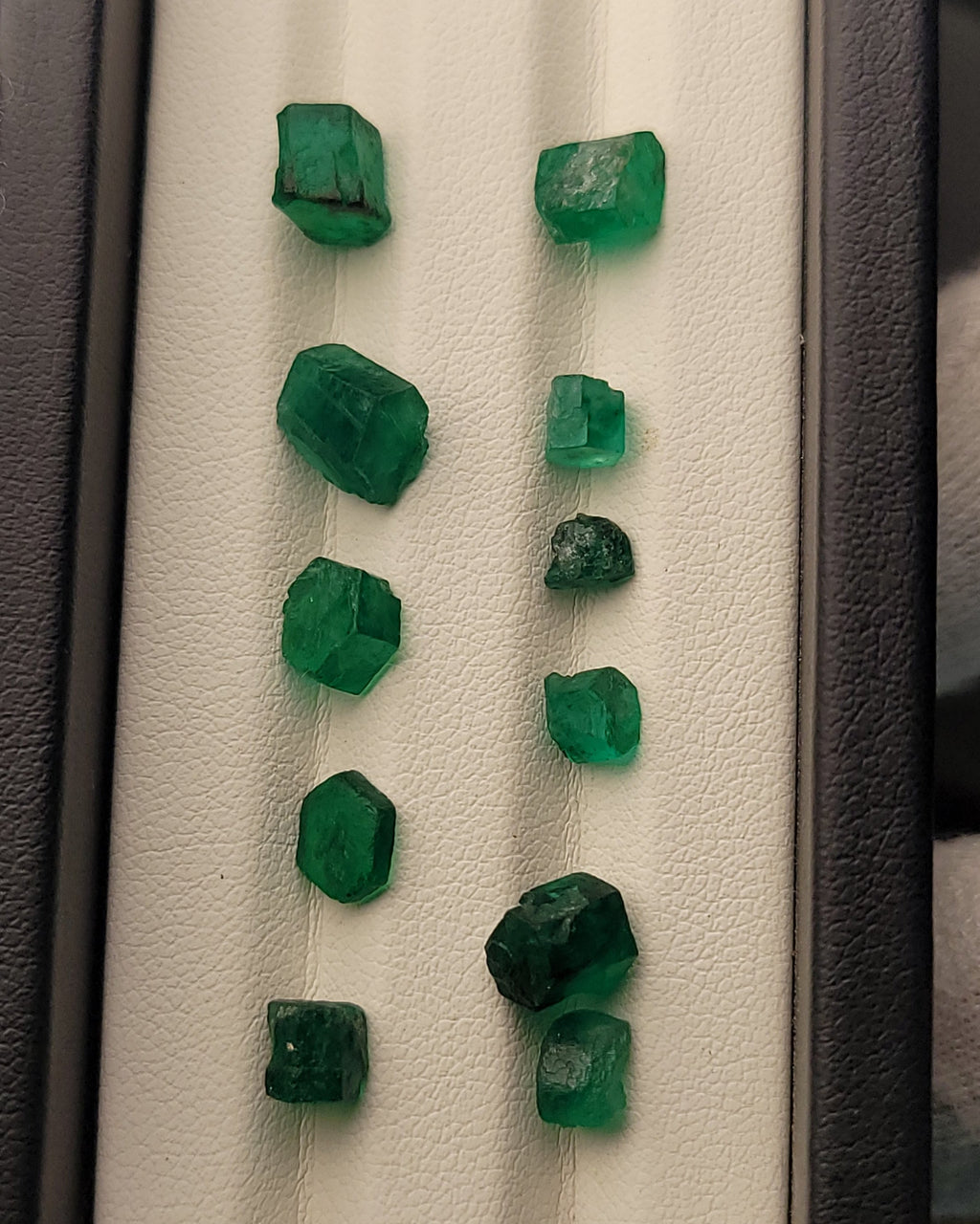 Nine raw green emerald crystals of varying sizes arranged in two columns on a white textured display card inside a dark gray jewelry box.