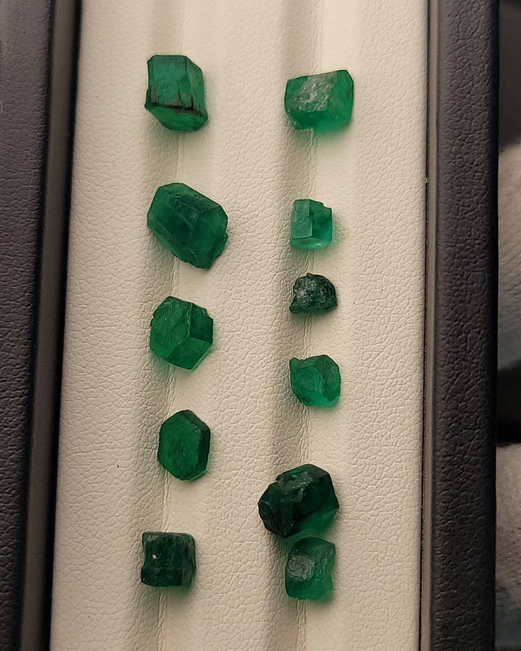 Nine raw green emerald crystals of varying sizes arranged in two columns on a white textured display card inside a dark gray jewelry box.