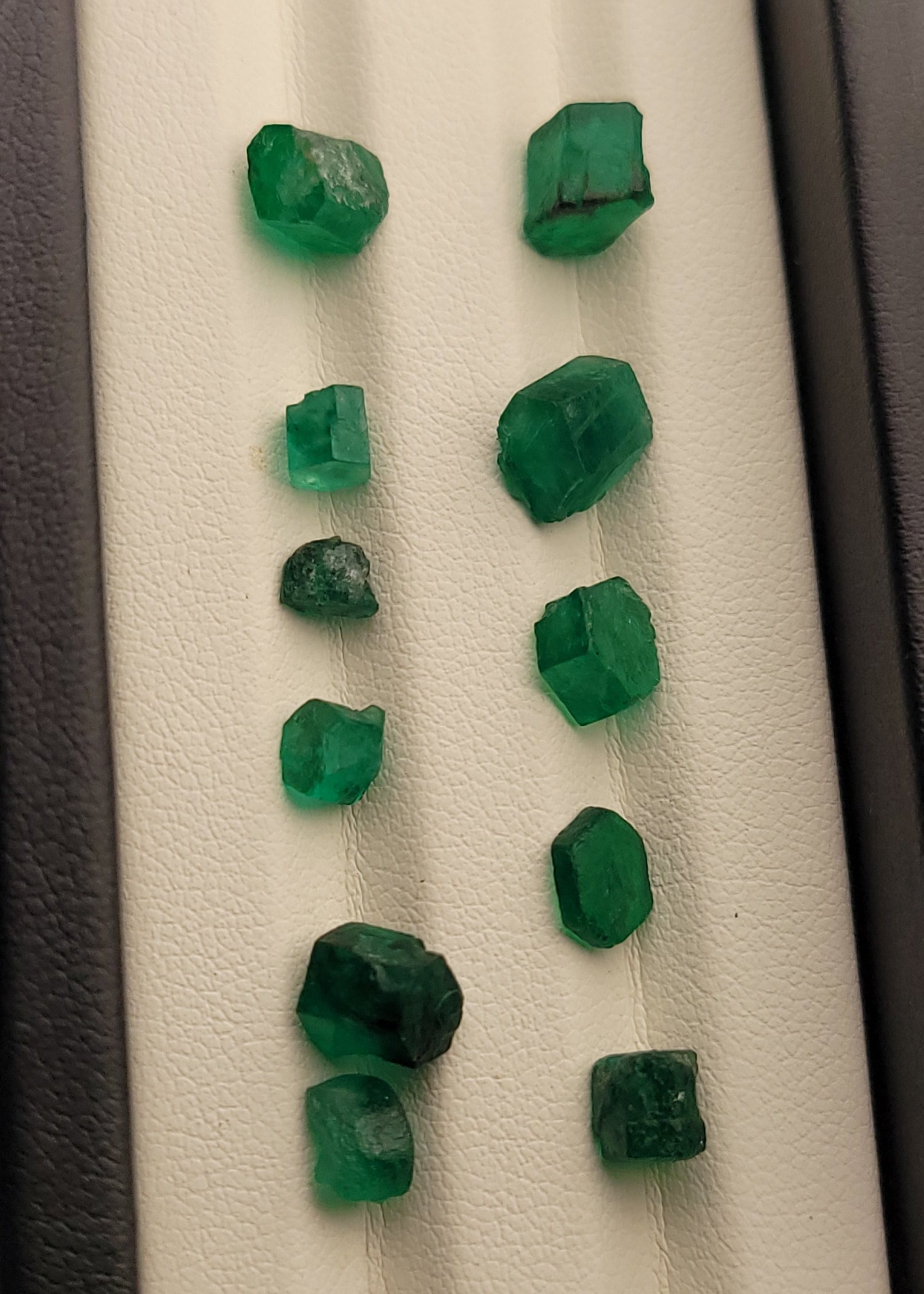 A set of rough, faceted green emerald-cut gemstones displayed in two vertical columns on a white textured surface inside a dark case.