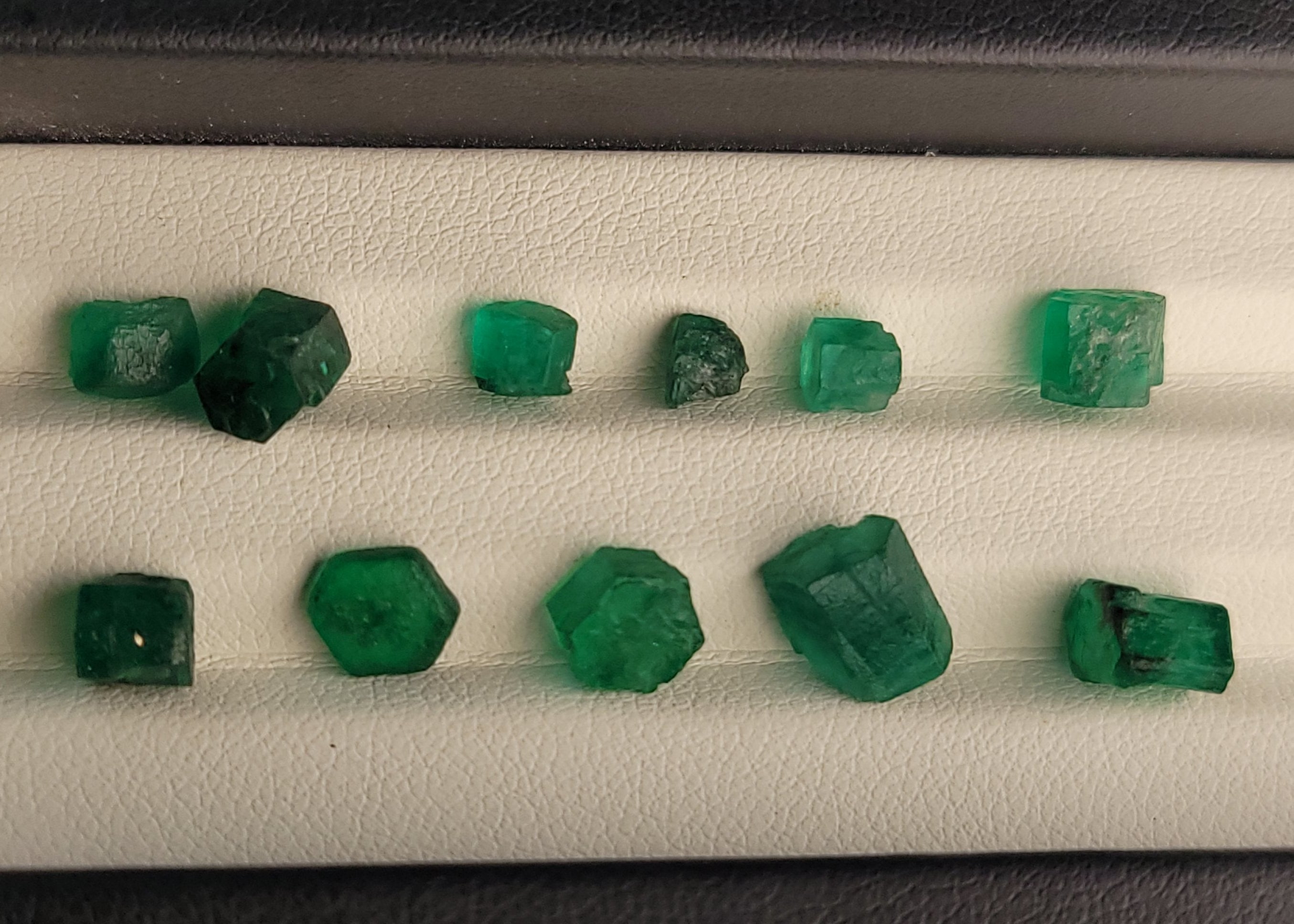 A set of rough, uncut green emerald-cut gemstones arranged in two rows on a white textured display tray inside a black case.