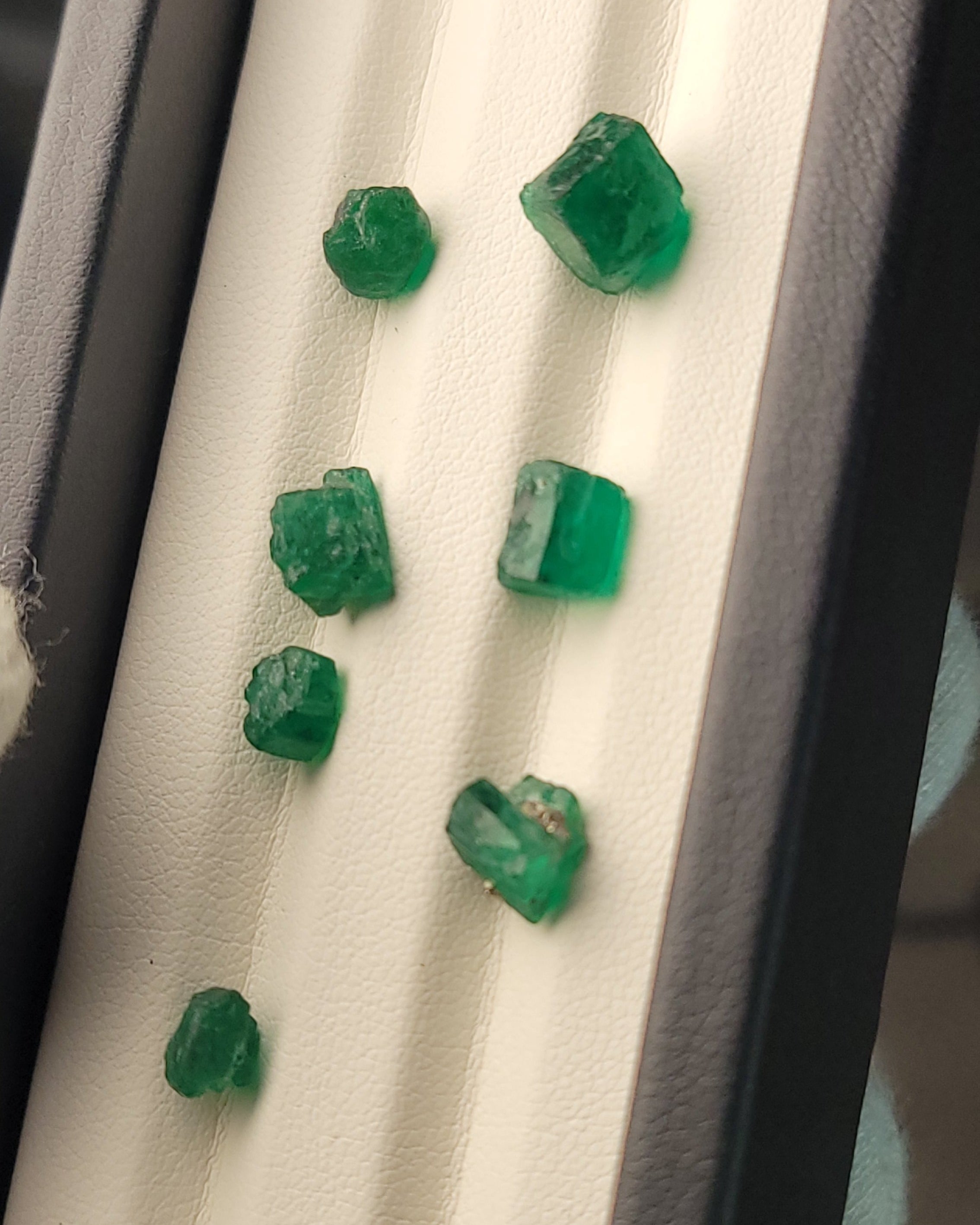 Several raw green emerald stones displayed on a white padded jewelry tray inside a dark case.