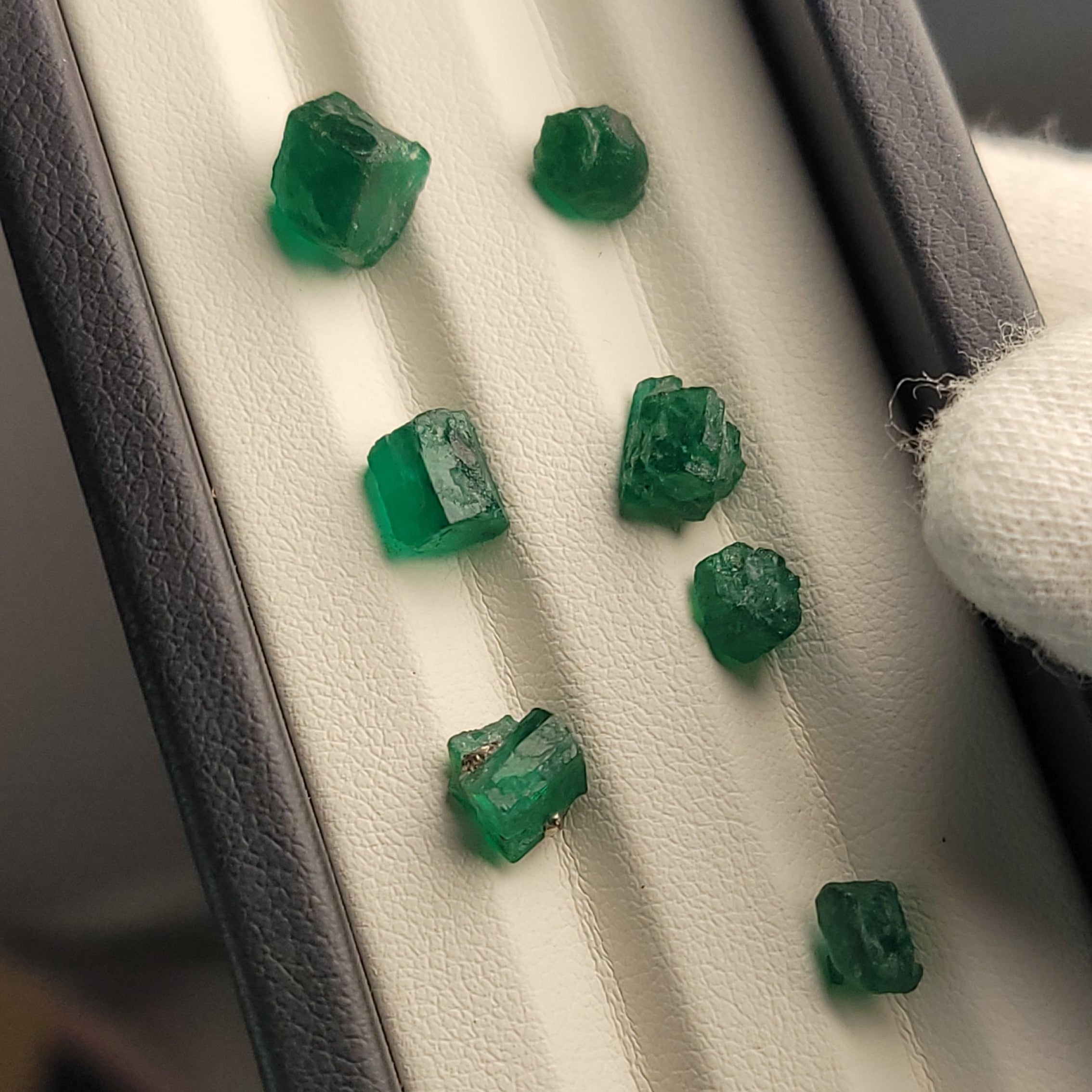 Several small green gemstone cubes arranged on a white padded tray inside a dark box, with a white gloved hand partially visible at the left edge.
