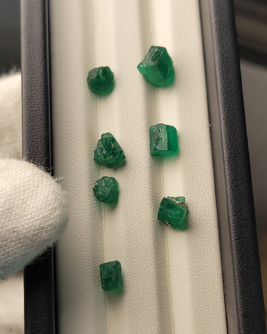 Seven raw, rough-cut green emeralds displayed on a white textured velvet insert inside a black presentation box, held by a white-gloved hand on the left.