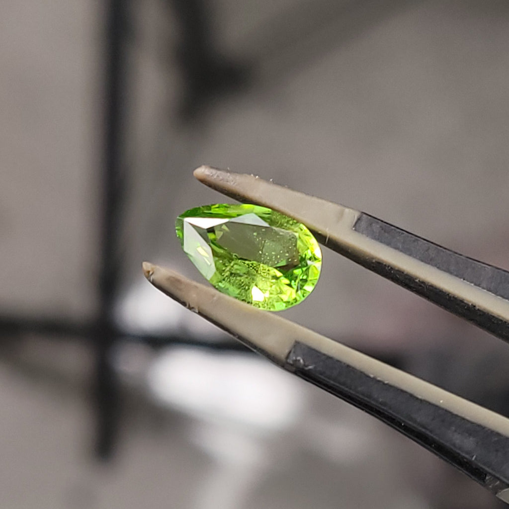 A small bright green faceted gemstone held in metal tweezers against a blurred indoor background.