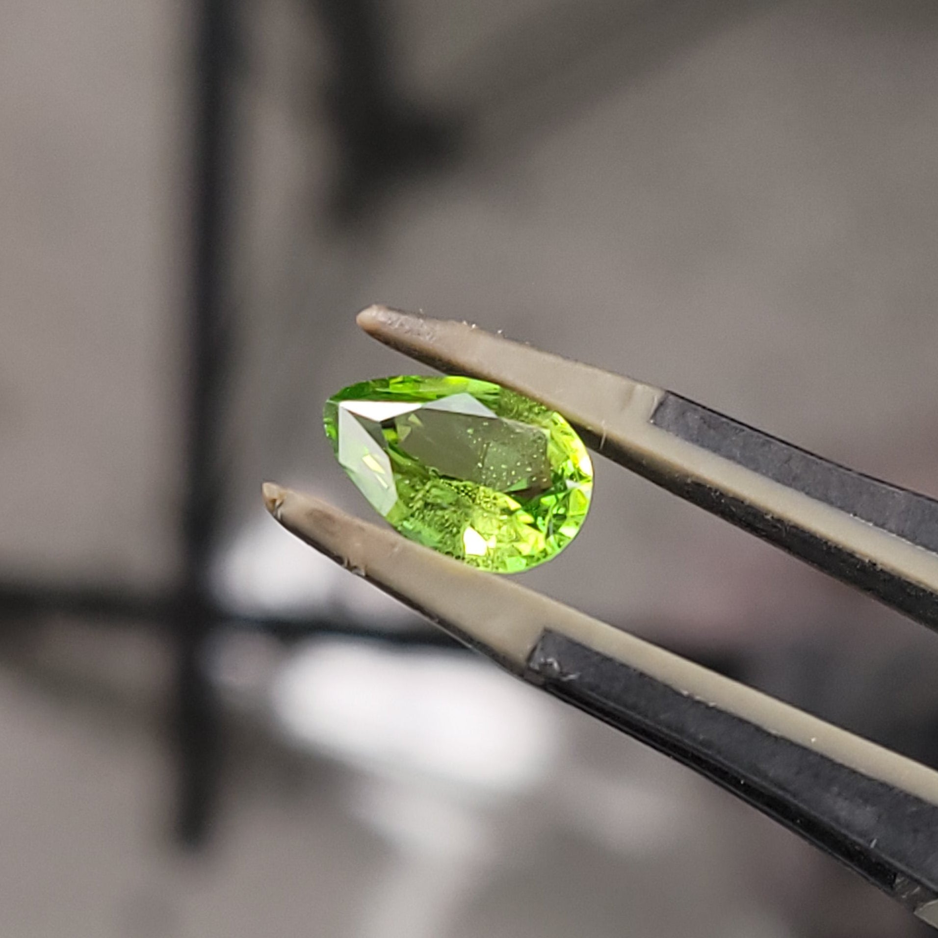 A small bright green faceted gemstone held in metal tweezers against a blurred indoor background.