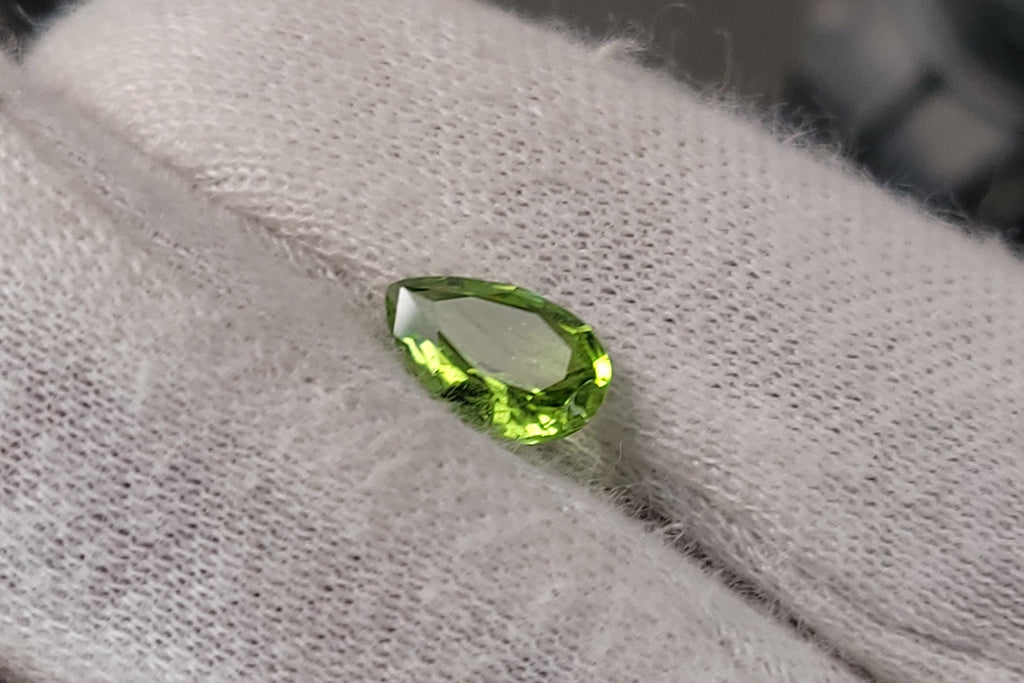 A small, faceted green gemstone, likely a peridot, resting on a white textured fabric surface.