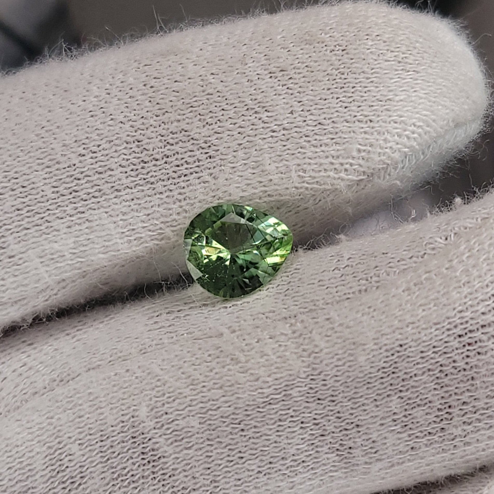A small round green gemstone held between white gloved fingers.